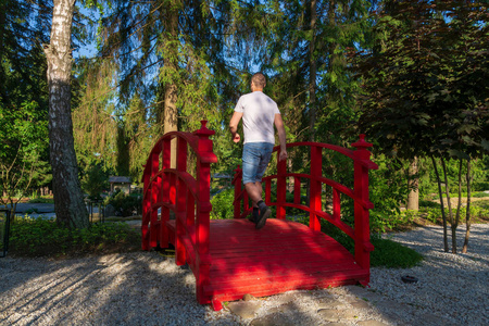 个穿着牛仔短裤和一件浅色t恤的年轻人走过公园里一座红色的木桥照片