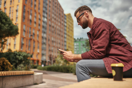 穿着休闲服留着胡茬的帅哥坐在户外的长椅上一边拿着手机一边喝着咖啡