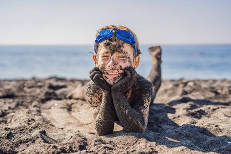 一个满脸脏兮兮的笑脸男孩在海里游泳前,坐在黑沙滩上玩耍.