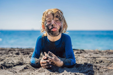 一个满脸脏兮兮的笑脸男孩在海里游泳前,坐在黑沙滩上玩耍.