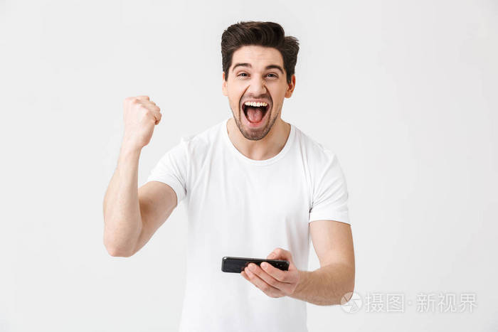 兴奋快乐情绪激动的年轻人摆出孤立的白墙背景玩游戏用手机做胜利者手势。