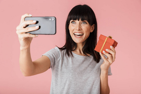 兴奋快乐女子用手机在浅粉色背景墙上摆出孤立的姿势拿着礼物盒自拍