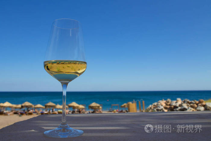 桌上放着一杯白葡萄酒里面有海滩的影子和倒影背景是天空和大海