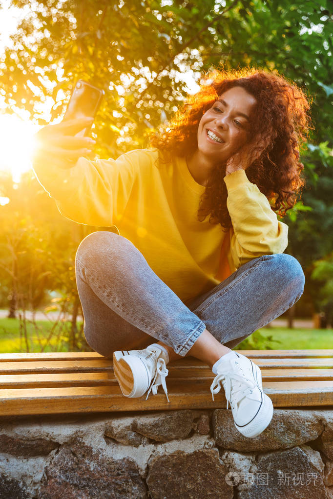 图为一位快乐的年轻女子坐在绿色公园的长椅上微笑着用智能手机自拍