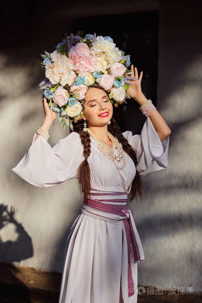 一个黑发女孩穿着白色的乌克兰正宗民族服装戴着花环对着一间白色小屋