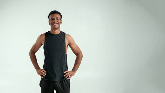 年轻英俊的美国黑人男子,双手叉腰,面带微笑地站在灰色背景下看着摄像