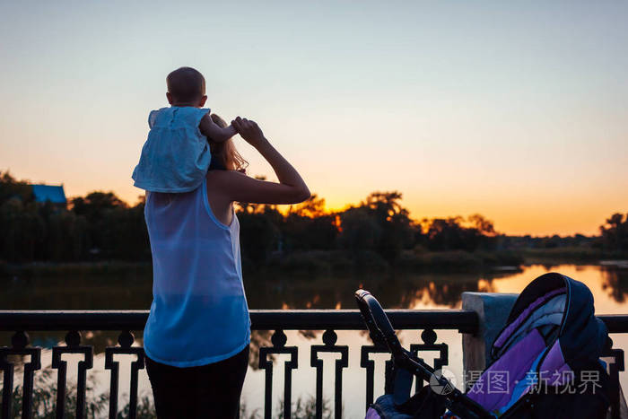 小女孩坐在妈妈的肩膀上欣赏风景夕阳下年轻的母亲和孩子在河边散步