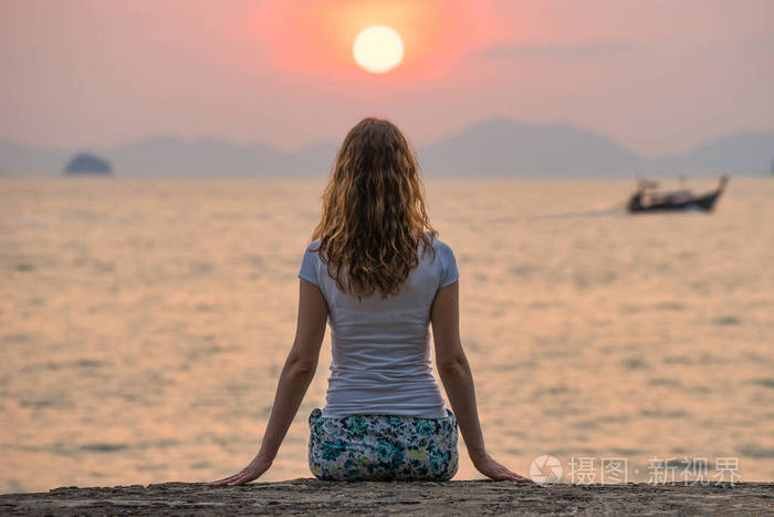 年轻女子独自坐在海边看着美丽的猩红夕阳