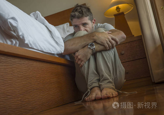 年轻的沮丧绝望的男人坐在卧室的地板上挨着床饱受抑