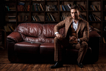 大胡子的商人坐在图书馆的豪华皮沙发上,喝着白兰地照片