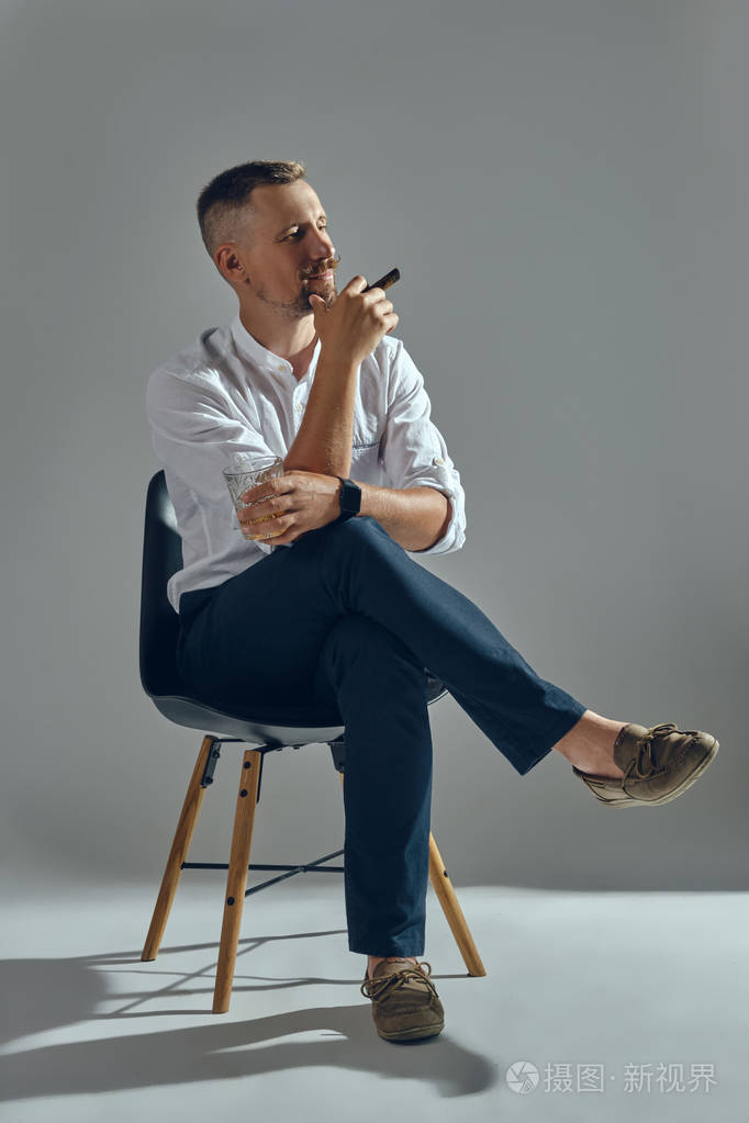 一个留着时髦胡子的男人穿着一件经典的白色衬衫和深色裤子坐在椅子上