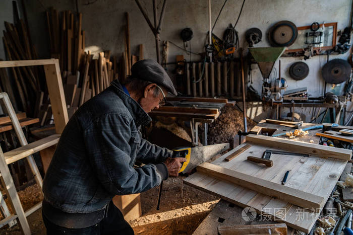戴着眼镜和灰色衣服的老木匠师傅在家里的车间里辛勤工作锯木板