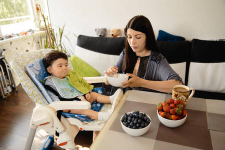 母亲在儿童座椅上喂宝宝吃粥-亚裔混血儿男童身穿蓝色体恤,笑脸熊卡通