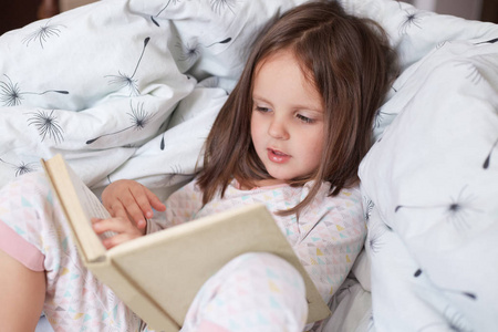 可爱的女孩在读她最喜欢的童话,孩子坐在床上看书,孩子准备上床睡觉