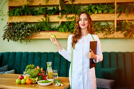 在绿色办公室里,身着医用制服的女营养师拿着苹果和不同的健康食品