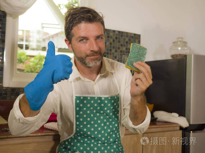 迷人的男人在厨房围裙里拿着洗碗海绵看起来积极愉快地享受家务劳动