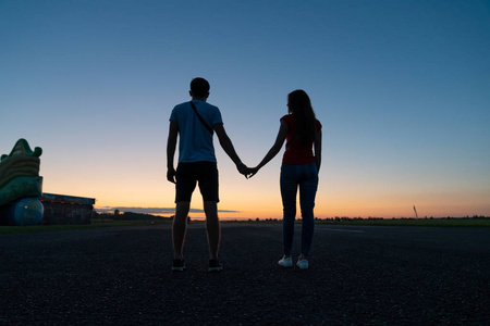 情侣站在一起背对着镜头,手牵手,遥望远方,美丽的夕阳映衬在背景上