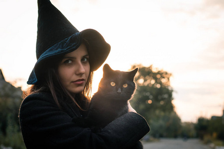 万圣节的猫在手和巫婆女孩戴着黑色帽子和外套在秋天的日落.