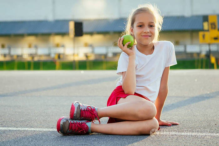 年轻漂亮的金发小女孩穿着运动服,白色t恤和红色运动短裤坐在沥青路上