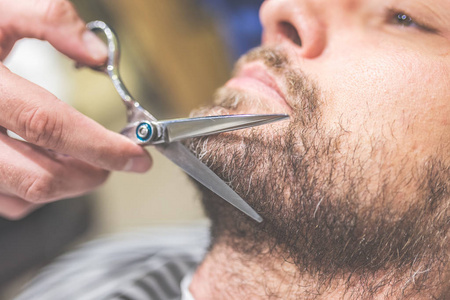 坐在理发店椅子上的年轻胡须男被理发师剪胡子的侧视图照片