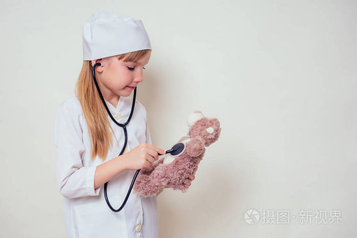 可爱的小女孩穿制服戴着医生的帽子和眼镜在白色的听诊器上玩医生和