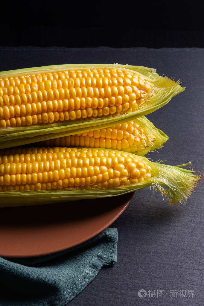 三个玉米穗绿色的叶子和盘子在黑暗的背景上