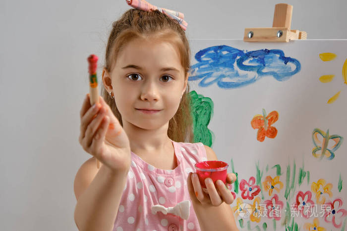 一个穿着粉红色裙子的小女孩站在画架后面用画笔在画布上画画在白色的