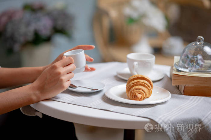 女性手拿咖啡杯的侧视图。在法国咖啡馆喝咖啡的女人。在法式餐厅喝咖啡的女人。白桌子上放羊角面包和咖啡。