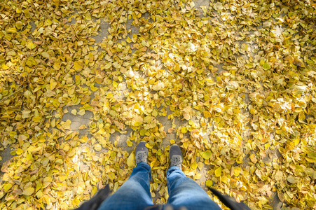 穿着蓝色牛仔裤和灰色运动鞋的男式脚踩在秋天的沥青上,上面有黄色的