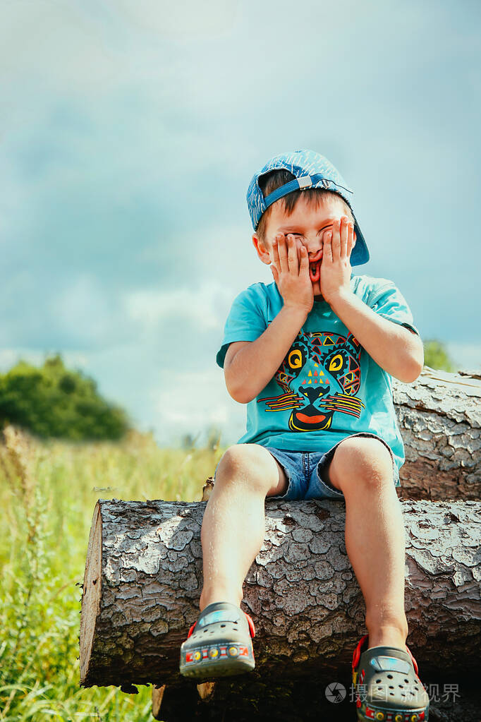 一个戴着棒球帽和牛仔短裤的学龄前儿童坐在圆木上嬉笑