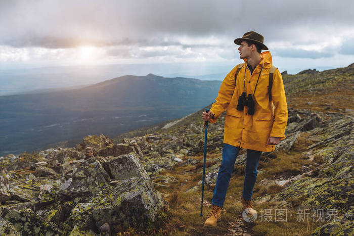 年轻的徒步旅行者，戴着帽子，穿着黄色外套，双筒望远镜站在岩石上看一边