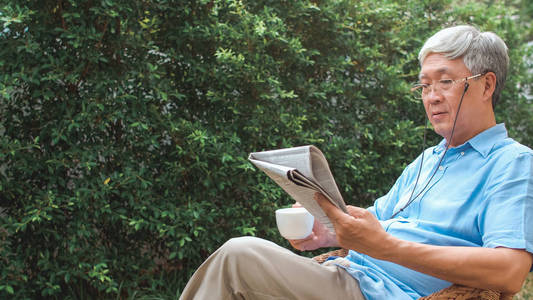 亚洲高级中国男性享受休息时间戴眼镜看报纸喝咖啡,而躺在花园里的家