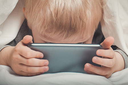 金发小男孩把鼻子埋在智能手机里.躺在床上,躲在被窝里.
