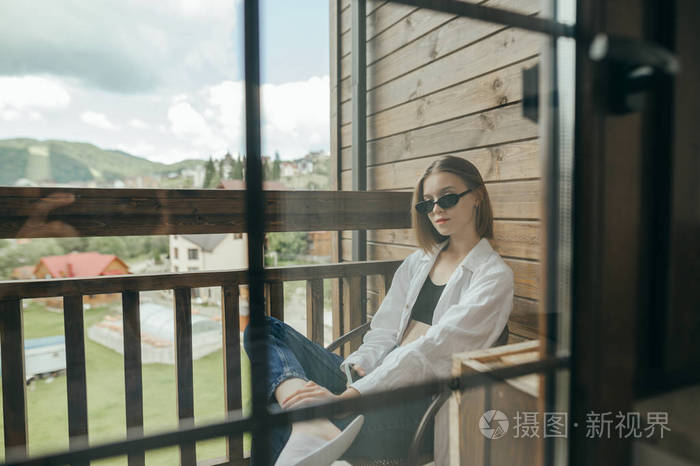 一个穿着休闲服戴着墨镜的女孩坐在酒店的阳台上看着镜头在乡村别墅