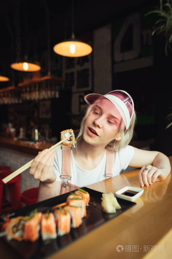 漂亮的女孩戴着粉红色的帽子穿着可爱的衣服拿着一卷寿司放在棍子上正