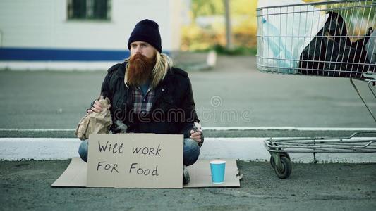 有胡须的年幼的无家可归的男人和卡纸板一次在近处购物