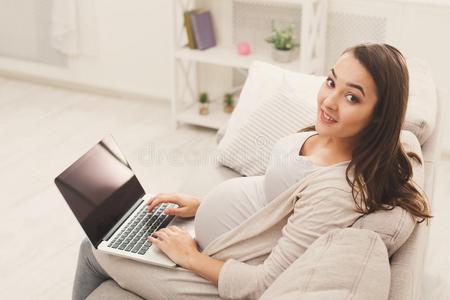 幸福的怀孕的女人使用便携式电脑在家