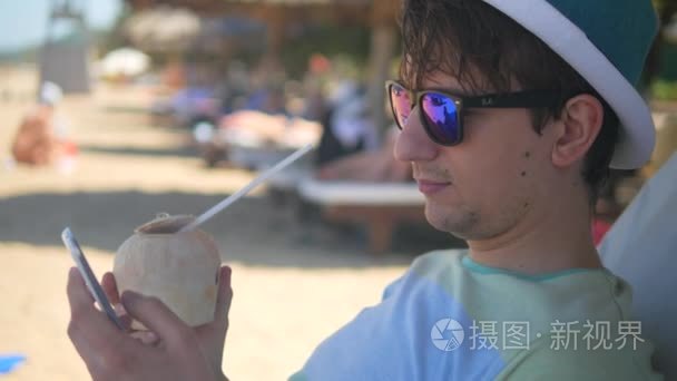 生活方式放松的人，使用手机和从一个在海滩上，手持拍摄的椰子喝一杯鸡尾酒