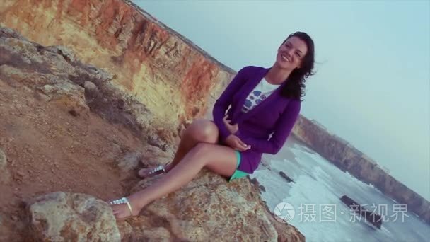 布鲁内特的年轻女孩，在紫色的外套，坐在岩石上在海上的短裤。夏天的傍晚。假期。笑