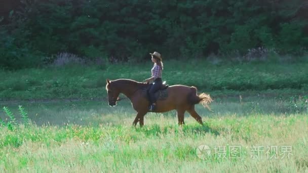 女孩戴着帽子在日落时分在慢动作骑一匹马在草地上的骑手