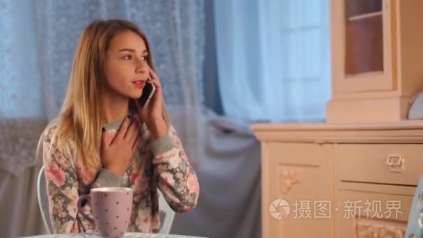 年轻美丽少女坐在桌前在客厅里说话的智能手机，和朋友，说长道短，喝茶聊天
