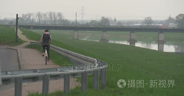 女人骑自行车沿着河景观绿色草坪谷城市景观汽车桥在光滑河车辆行驶在春天阴天