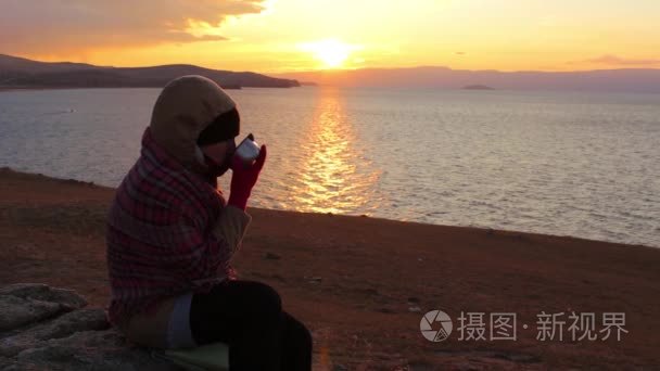 女孩坐在海边，喝热茶。令人惊异的金色的夕阳