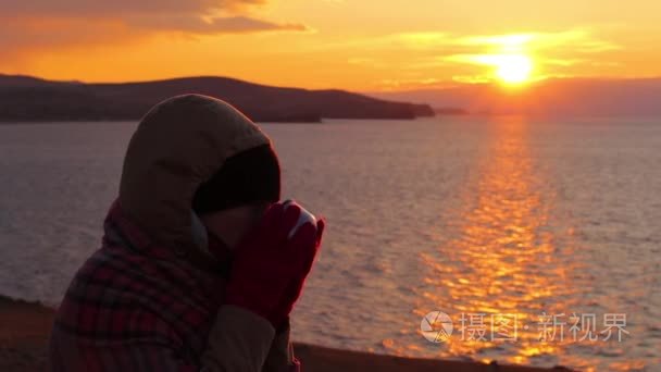 女孩坐在海边，喝热茶。美丽的金色的夕阳