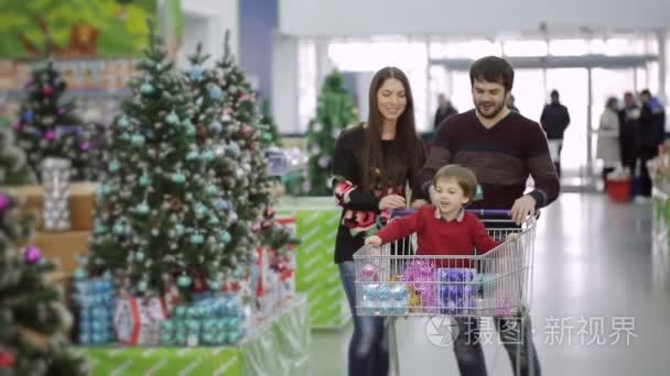 美好的家庭，走在大厅与超市购物车和选择圣诞节玩具和圣诞礼物。小男孩坐在购物车，并考虑购买