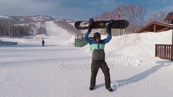 快乐男性滑雪板与他手中的滑雪板是站在滑雪胜地和快乐的舞蹈。在冬天的衣服的人是开心和山坡上上升与董事会的手