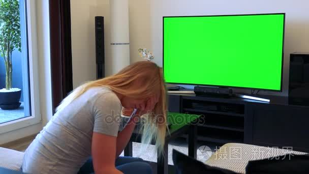 一个金发碧眼的女人坐在客厅里的沙发上看电视与绿色的屏幕，然后不幸的是用双手蒙住脸视频