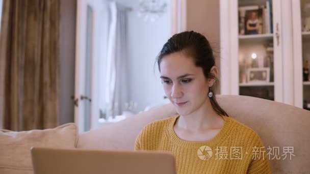年轻的女人坐在客厅沙发上使用笔记本电脑。女性在家里工作，上网冲浪