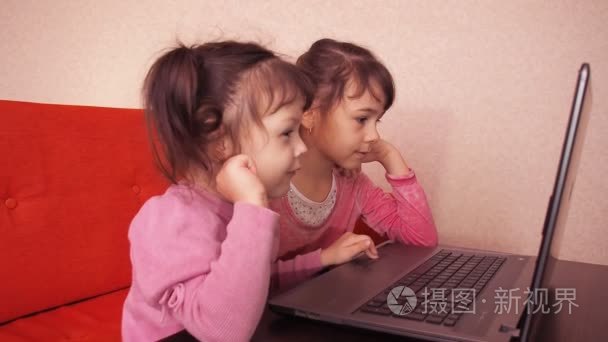 孩子们玩在便携式电脑中。两个小女孩在一台笔记本电脑上打印。两姐妹坐在橙色沙发上，看着一台笔记本电脑