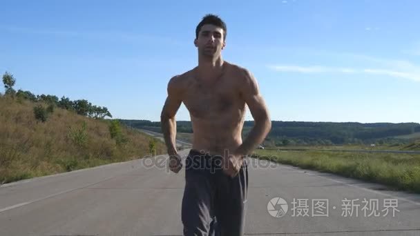 在公路慢跑的短跑运动员男人。男运动员在夏季训练户外。年轻强壮肌肉的人在锻炼期间在农村公路上行使。外积极健康的生活方式。关闭